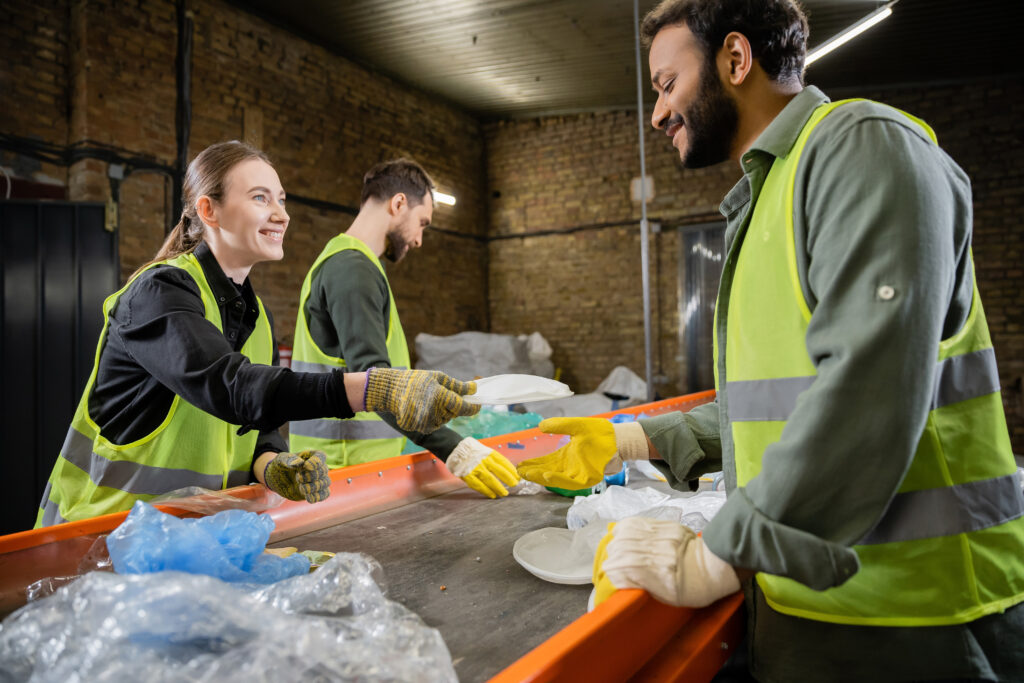 cheerful sorter in high visibility vest and gloves giving plastic trash to indian colleague while working together near conveyor in waste disposal station, garbage sorting and recycling concept